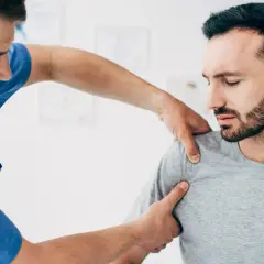 Fizjoterapeuta bada pacjenta z podejrzeniem zwichniętego barku. Mężczyzna w niebieskim fartuchu delikatnie uciska ramię pacjenta, który ma zamknięte oczy i grymas bólu.