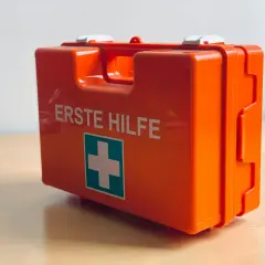 Pomarańczowa apteczka z napisem "ERSTE HILFE" i zielonym krzyżem.