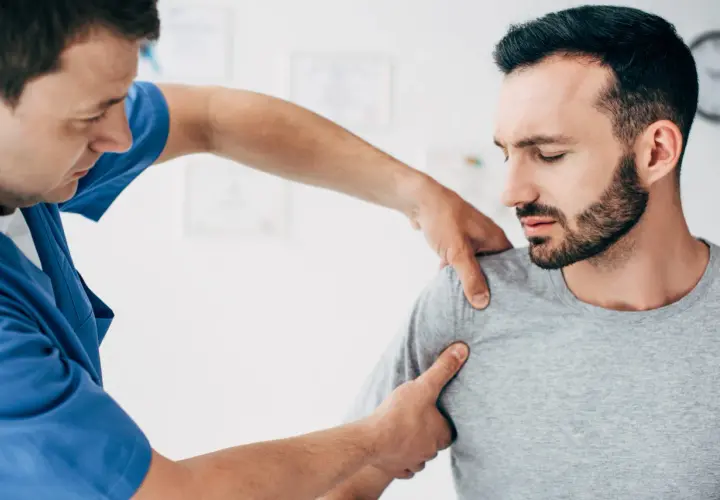 Fizjoterapeuta bada pacjenta z podejrzeniem zwichniętego barku. Mężczyzna w niebieskim fartuchu delikatnie uciska ramię pacjenta, który ma zamknięte oczy i grymas bólu.
