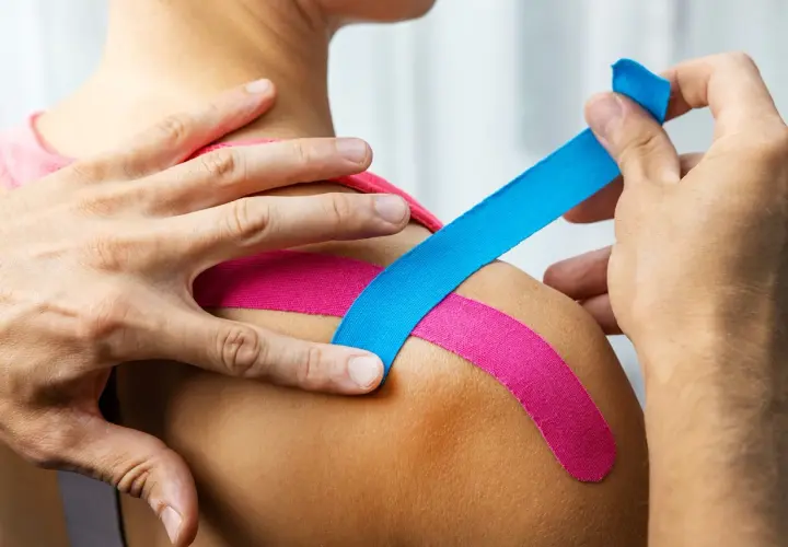 Dłonie aplikują kolorowy plaster kinesiotaping na ramię, wspierając mięśnie i stawy.