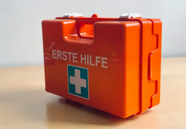 Pomarańczowa apteczka z napisem "ERSTE HILFE" i zielonym krzyżem.