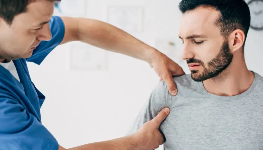 Fizjoterapeuta bada pacjenta z podejrzeniem zwichniętego barku. Mężczyzna w niebieskim fartuchu delikatnie uciska ramię pacjenta, który ma zamknięte oczy i grymas bólu.