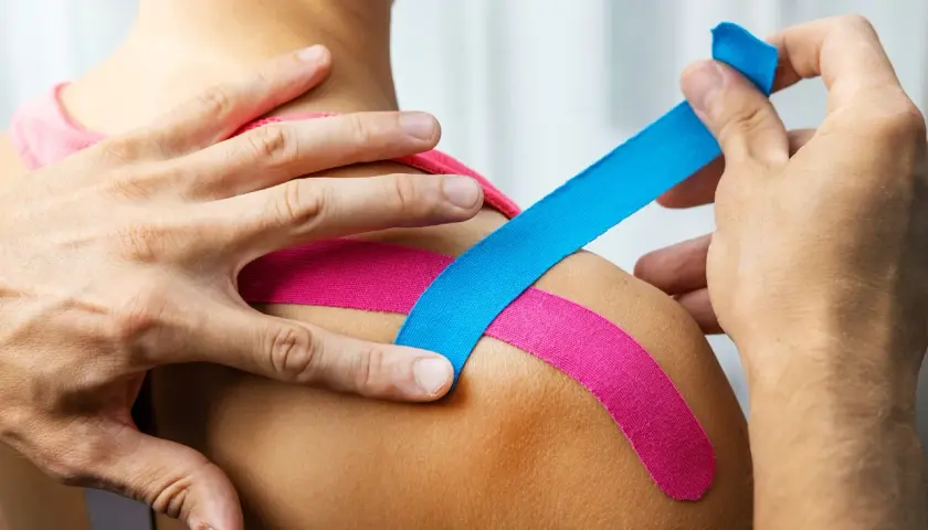 Dłonie aplikują kolorowy plaster kinesiotaping na ramię, wspierając mięśnie i stawy.