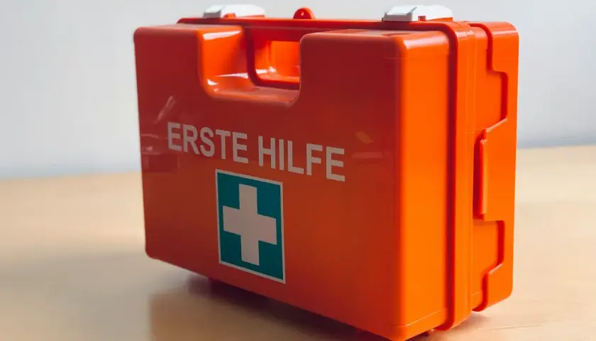Pomarańczowa apteczka z napisem "ERSTE HILFE" i zielonym krzyżem.