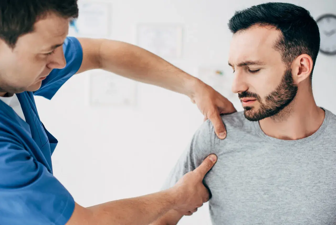 Fizjoterapeuta bada pacjenta z podejrzeniem zwichniętego barku. Mężczyzna w niebieskim fartuchu delikatnie uciska ramię pacjenta, który ma zamknięte oczy i grymas bólu.