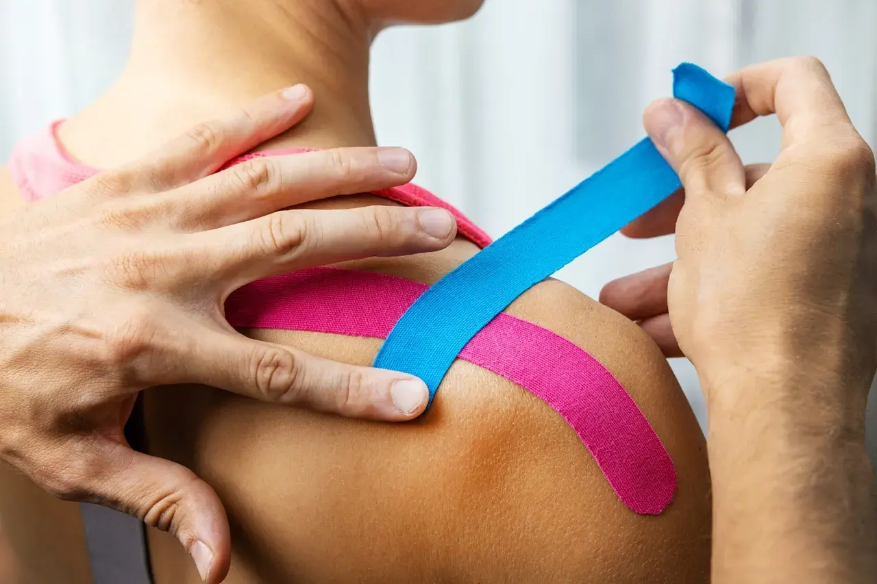 Dłonie aplikują kolorowy plaster kinesiotaping na ramię, wspierając mięśnie i stawy.