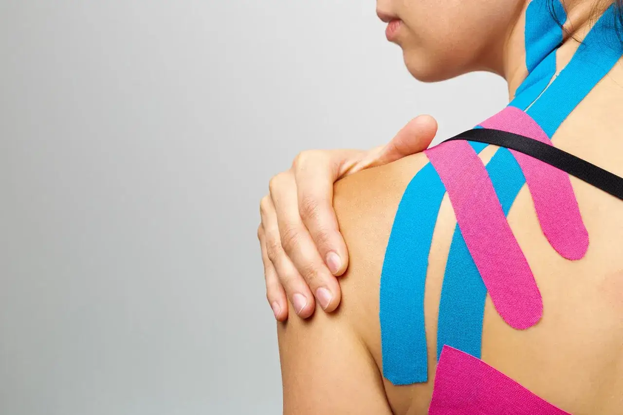 Plaster kinesiotaping w kolorach niebieskim i różowym na ramieniu i plecach kobiety. Dłoń spoczywa na ramieniu.